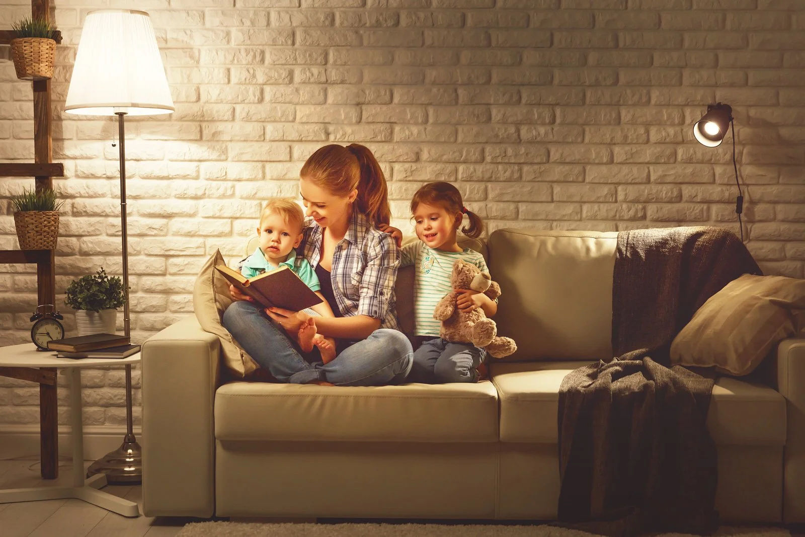 A woman sits on a sofa in Orange, California, reading a book to two young children; one holds a teddy bear. A lamp and blankets are visible in the cozy, softly lit room.