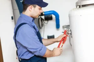 A plumber in a blue uniform and cap uses a wrench to adjust a pipe on a white water heater, performing water heater maintenance in Brea, California.