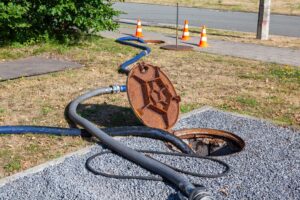 Open manhole with hoses inserted, possibly indicating a hidden pipe leak, surrounded by gravel and grass, with traffic cones placed nearby on a sunny day.