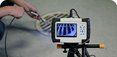 A person uses a video inspection camera to examine the inside of a drain through its metal grate cover.