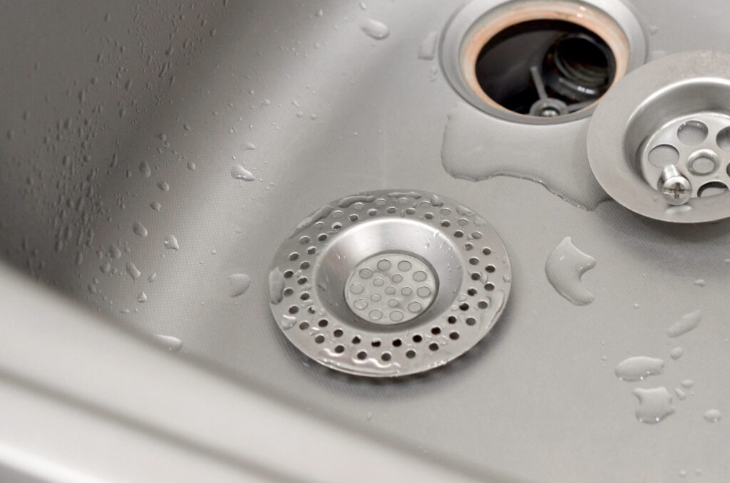 A close-up of a stainless steel kitchen sink with water droplets, highlighting a metal sink strainer and drain stopper—helpful for preventing clogged drains and common plumbing problems.