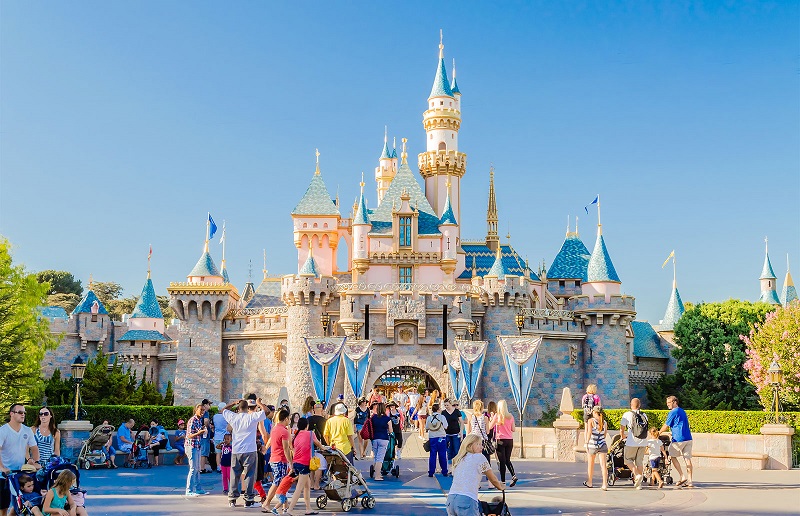 People walk and gather in front of the Sleeping Beauty Castle at Disneyland in Orange County, CA, on a sunny day, with clear blue sky and trees in the background.