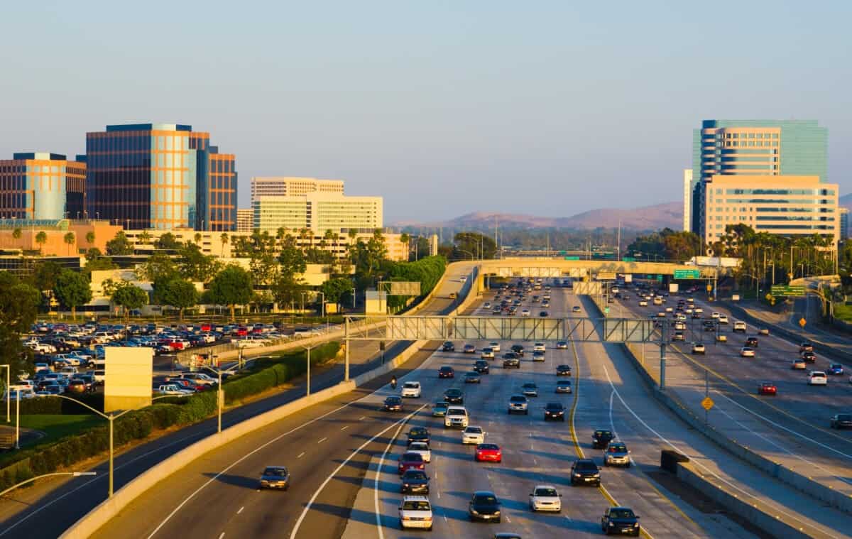 A busy freeway with multiple lanes of cars and light traffic moves through an Orange County CA city area, with office buildings and a pedestrian bridge in the background.