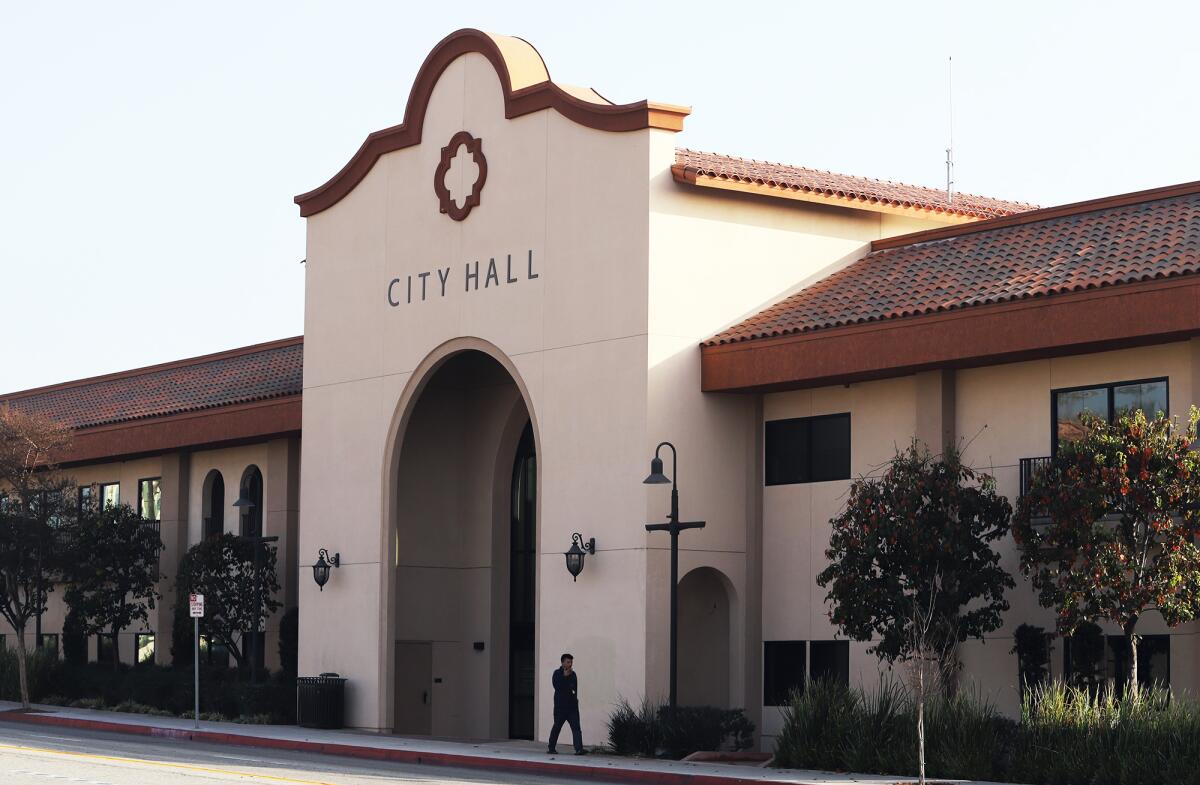 A person walks in front of a beige building with an arched entrance labeled "City Hall," nestled among trees in Orange County, California.