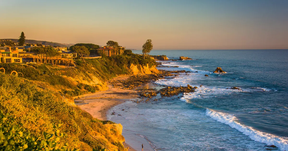 A coastal scene in Orange County, CA shows rocky cliffs, a sandy beach, ocean waves, and houses overlooking the shoreline at sunset.