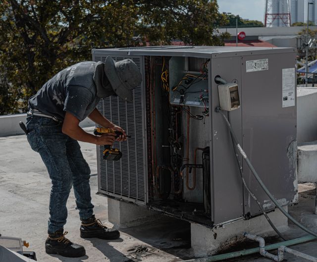A person wearing a hat and work boots uses a drill to repair or inspect an outdoor air conditioning unit on a rooftop.