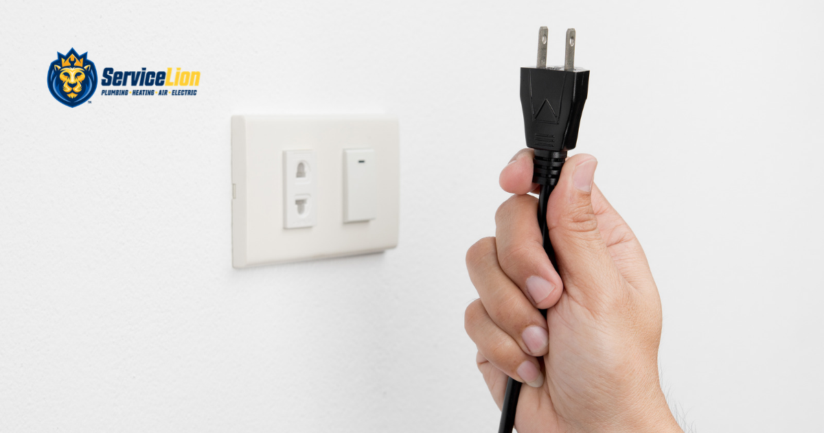 A hand holds a black electrical plug near a wall outlet and switch, highlighting the importance of electrical safety. The ServiceLion plumbing, network, and electrical logo is visible in the top left corner.