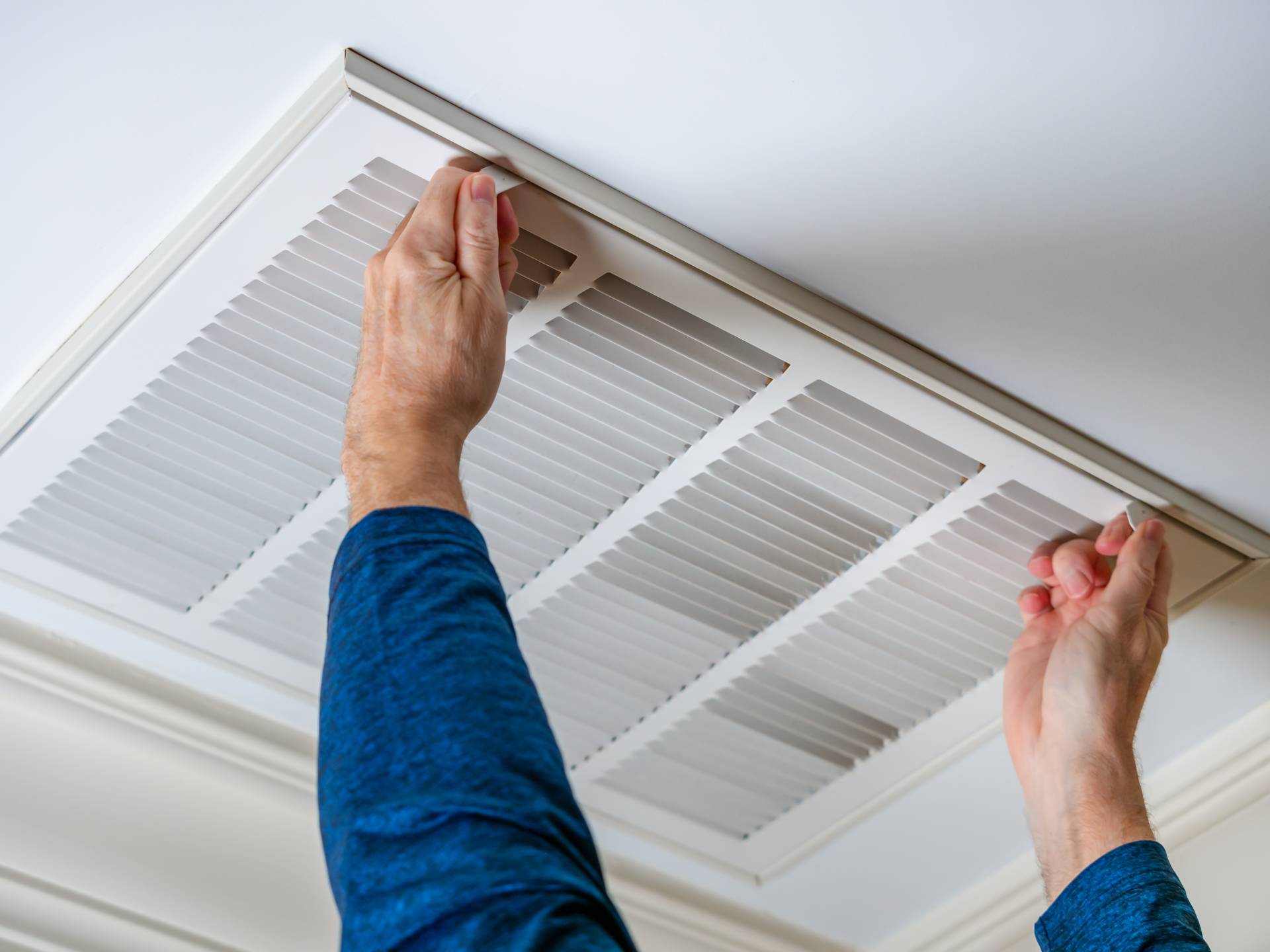 A person in a blue shirt removes a rectangular air vent cover from a white ceiling.