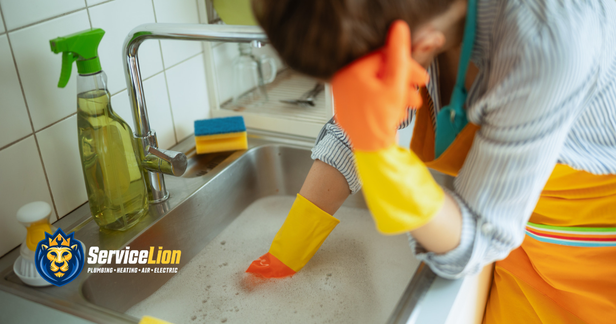 Person in yellow gloves leans on kitchen sink filled with soapy water, looking tired from kitchen sink backup. ServiceLion Plumbing-Heating-Air-Electric logo appears in the lower left corner.