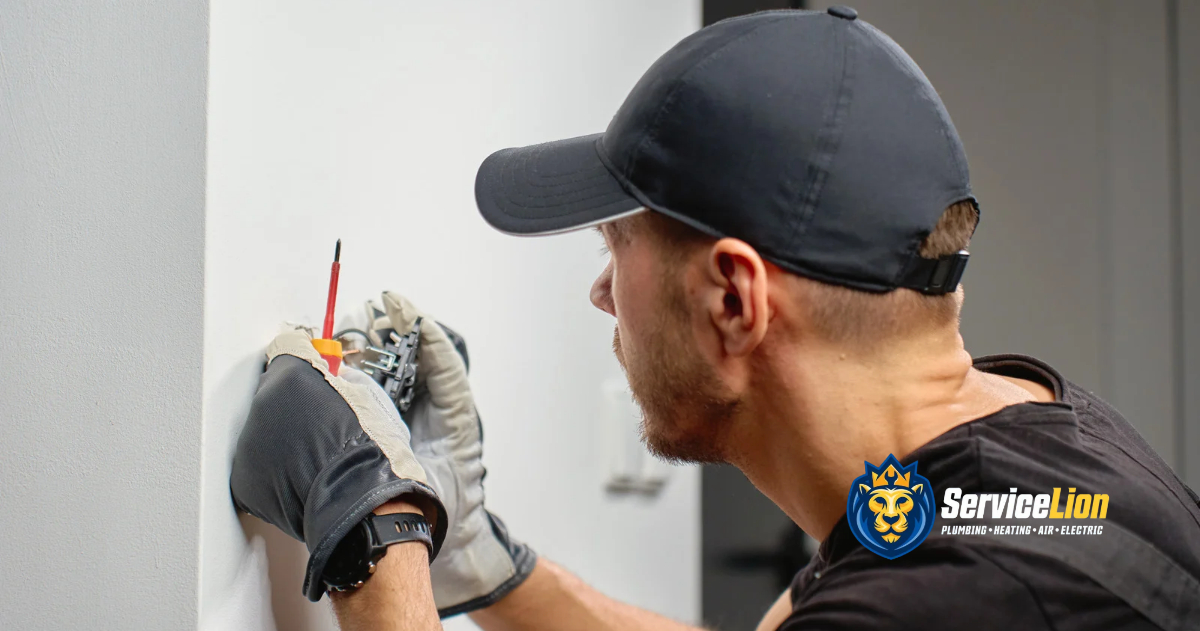 A technician in a black cap and work gloves uses a screwdriver to repair a buzzing electrical outlet.