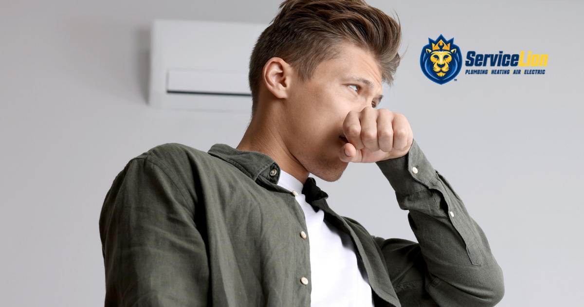 A person covers their nose and mouth near a wall-mounted air conditioner emitting a musty AC smell, with the ServiceLion logo in the corner.