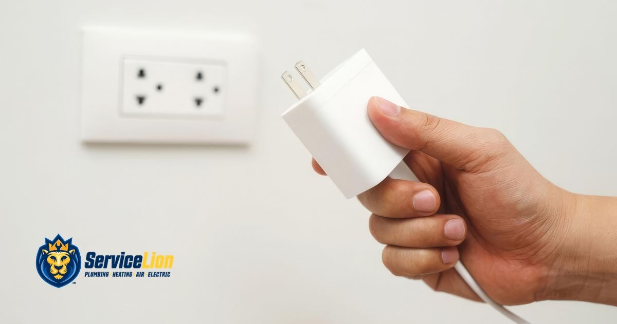 A hand holding a white power adapter near a wall outlet, with the ServiceLion logo in the bottom corner.